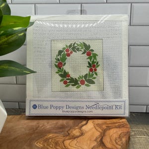 Puede incluir: Un kit de punto de cruz con un diseño de corona de hojas verdes y bayas rojas. El kit está en un marco blanco, con el texto "Blue Poppy Designs Needlepoint Kit" y la dirección del sitio web.
