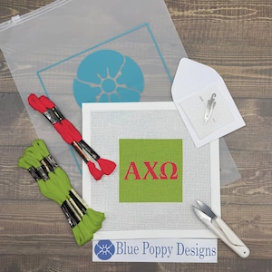 May include: A needlepoint kit with a white canvas featuring a green square with the Greek letters AXΩ in red. Includes red and green embroidery floss, a needle, scissors, and a clear plastic bag with a blue design.
