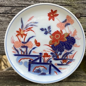 May include: A white ceramic plate with a blue and red floral design. The flowers are stylized and have gold accents. The plate has a raised rim.