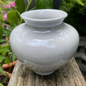 May include: A small, round, light gray ceramic vase with a wide opening and a slightly flared rim. The vase has a smooth, glossy finish and sits on a wooden surface. Green foliage is visible in the background.