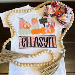 May include: White t-shirt with a colourful pumpkin patch design. The shirt features the name "ELLASYN" in large black letters. A matching bow with the name "ELLASYN" is also visible. A wooden bead garland with tassels frames the shirt.