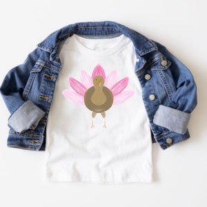 Può includere: T-shirt bianco con un'illustrazione di un tacchino dei cartoni animati. Il tacchino ha piume rosa e un corpo marrone. Una giacca di jeans blu è drappeggiata sulla maglietta, con le maniche arrotolate.