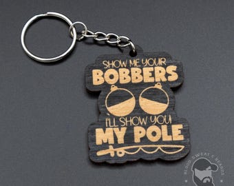 Könnte beinhalten: Dunkler Holzschlüsselanhänger mit silbernem Schlüsselring. Der Schlüsselanhänger trägt den Text "SHOW ME YOUR BOBBERS I'LL SHOW YOU MY POLE" mit Grafiken von Angelposen. Ein Logo ist unten rechts zu sehen.