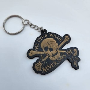 May include: A black wooden keychain with a gold skull and crossbones design. The skull has one eye and is wearing a hat. The text "One Eyed Willie Never Says Die" is written on a banner.