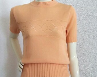 May include: A peach-colored short-sleeved sweater with a diamond pattern on the front. The sweater is paired with a white pleated skirt.