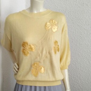 May include: A pale yellow sweater with short sleeves and a crew neck. The sweater has a floral design with gold embroidered flowers.