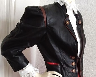 May include: A black leather jacket with brown trim and gold buttons. The jacket has a stand-up collar and a fitted silhouette. The model is wearing a white lace blouse and a red pleated skirt.