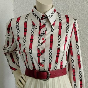 Może przedstawiać: Strój w stylu vintage na manekinie. Zestaw obejmuje białą bluzkę z czerwono-czarnym wzorem łańcucha, czerwony pasek i kremową plisowaną spódnicę. Bluzka ma długie rękawy i kołnierzyk.