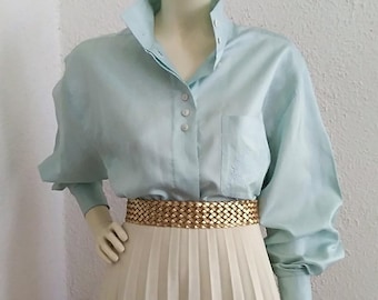May include: A light blue button-down shirt with long sleeves and a white pleated skirt. The shirt has a collar and a pocket on the left side. The skirt has a gold woven belt.