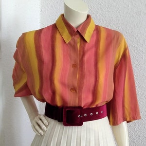 Puede incluir: Una camisa de manga corta a rayas con cuello y cierre de botones. La camisa es en tonos de rosa, naranja y amarillo. Un cinturón burdeos se lleva alrededor de la cintura.