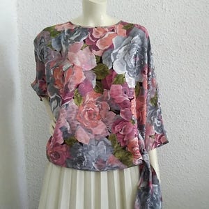 Puede incluir: Una blusa con estampado floral con detalle de lazo, combinada con una falda plisada. La blusa presenta un estampado de rosas rosas, grises y verdes. La falda es de color crema claro.