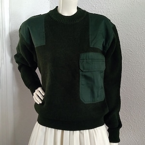 Może przedstawiać: Zielony sweter z dużą naszywaną kieszenią po prawej stronie. Sweter ma żebrowaną fakturę i okrągły dekolt.