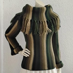 Op de afbeelding: Een gestreepte trui in groen, bruin en beige, getoond op een mannequin. De trui heeft een hoge kraag en franjes op de schouders. De mouwen zijn uitlopend en gecombineerd met een witte rok. Afmetingen in centimeters.