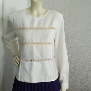 Può includere: Blusa bianca a maniche lunghe con un davanti plissettato e tre file di pizzo. La blusa è indossata sopra una gonna plissettata viola.
