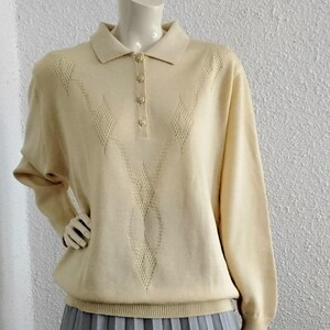 Può includere: Un maglione color crema a maniche lunghe con collo e tre bottoni. Il maglione presenta un motivo traforato a forma di diamante sul davanti. Abbinato a una gonna plissettata grigia, l'outfit è esposto su un manichino.