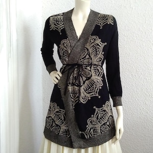 Op de afbeelding: Een zwarte en gouden cardigan met een wikkelmodel en een bloemenpatroon. De cardigan heeft een strikceintuur en lange mouwen.