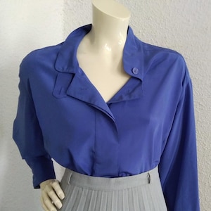 Pode incluir: Uma blusa azul de manga comprida com colarinho e fecho de botões. A blusa é usada sobre uma saia plissada cinzenta.