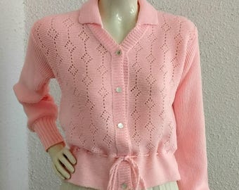 May include: A pale pink cardigan with a collared neckline and long sleeves. The sweater has a diamond-patterned openwork design and a drawstring waist. It is buttoned and displayed on a mannequin.