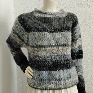 May include: A hand-knitted sweater with horizontal stripes in shades of grey, black, and blue. The sweater has a crew neck and long sleeves. The garment is displayed on a mannequin, paired with a cream-coloured pleated skirt.