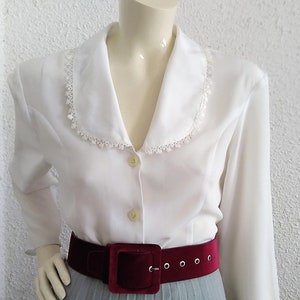 Peut inclure: Chemise blanche à manches longues avec un col en dentelle et une fermeture boutonnée. La chemise est portée avec une ceinture en velours bordeaux.