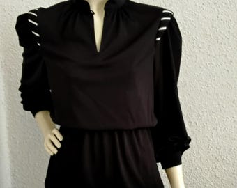 May include: Black long-sleeve blouse with a V-neck and white accent stripes on the shoulders. The blouse is paired with a gray pleated skirt. The mannequin is displayed against a white textured wall. The outfit is a vintage style.