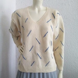 Può includere: Un maglione color crema con scollo a V e un motivo geometrico blu e bianco.