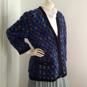 May include: A blue and black checkered cardigan with a black trim and a pocket on the left side. The cardigan is made of a soft, fuzzy material.