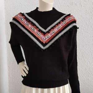 Peut inclure: Pull noir à col montant et manches longues. Le pull présente un motif en forme de V sur la poitrine avec des détails brodés dans les tons de rouge, orange et blanc. Le motif est orné de petites pompons noirs.