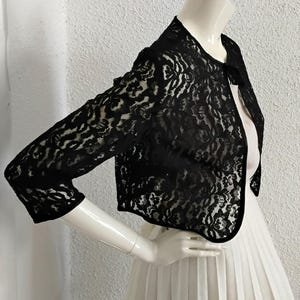 May include: Black lace bolero jacket with three-quarter sleeves. The jacket is displayed on a mannequin over a white dress. The lace has a floral pattern, and the jacket has a rounded neckline and a black trim.