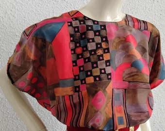 May include: A colorful, abstract-patterned blouse with short sleeves. The top features a mix of red, blue, brown, and orange geometric shapes. A red belt with a gold buckle cinches the waist. The blouse is paired with a white pleated skirt.