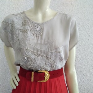 Può includere: Una blusa grigia chiaro con un delicato ricamo floreale. La blusa ha maniche corte e un collo rotondo. La blusa è abbinata a una gonna plissettata rossa e una cintura dorata.