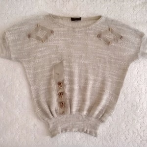 Könnte beinhalten: Ein beiger Strickpullover mit kurzen Ärmeln und geripptem Saum. Der Pullover hat zwei dekorative braune Stoffapplikationen an den Schultern und einen braunen Stoffstreifen mit Knöpfen an der Vorderseite.