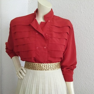 May include: A red blouse with layered detailing across the chest and a mandarin collar. The blouse is buttoned and has long sleeves. A gold woven belt cinches the waist, paired with a white pleated skirt.