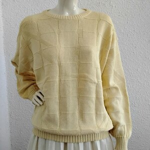 Pode incluir: Um suéter amarelo pálido com um padrão geométrico, exibido em um manequim. O suéter tem gola redonda, mangas compridas e punhos e bainha canelados. O manequim está usando uma saia plissada branca.