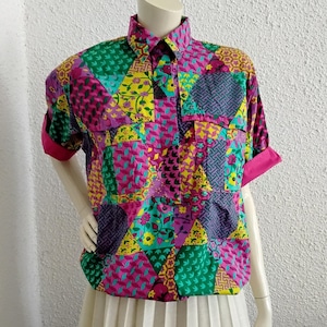 Peut inclure: Une chemise à manches courtes avec un motif patchwork dans les tons de rose, vert, jaune et violet. La chemise a un col, deux poches avant et des poignets roses. La chemise est présentée sur un mannequin avec une jupe plissée blanche.