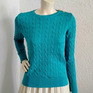 Peut inclure: Un pull en tricot torsadé de couleur turquoise, à manches longues et col rond. Le pull a des boutons dorés sur l'épaule et est présenté sur un mannequin. Le bas du mannequin porte une jupe plissée blanche.