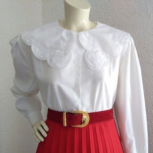 Puede incluir: Una blusa blanca con un cuello grande de encaje y mangas largas. La blusa se lleva con una falda plisada roja y un cintur&oacute;n dorado.