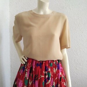 Könnte beinhalten: Eine beige Seidenbluse mit kurzen Ärmeln und einem runden Ausschnitt. Die Bluse wird mit einem roten Rock mit Blumenmuster kombiniert.
