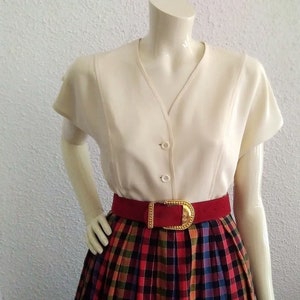 May include: A cream-coloured, short-sleeved blouse with a V-neck and button closure. A wide, red belt with a gold buckle cinches the waist above a plaid skirt in red, green, blue, and yellow. The outfit is displayed on a mannequin.
