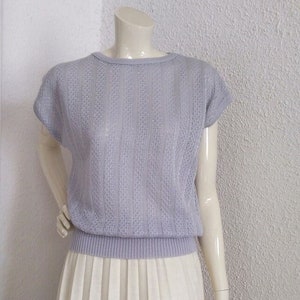 May include: A pale purple knitted top with short sleeves and a ribbed hem, combined with a long, pleated cream-coloured skirt. The top features a textured, open weave design. The outfit is displayed on a mannequin.