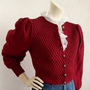 May include: A red knitted cardigan with puffed sleeves and a diamond pattern. The cardigan has a button closure and is worn over a white lace blouse.