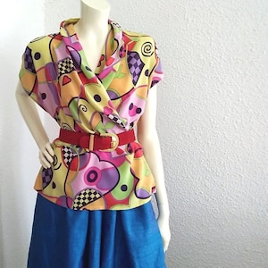 Könnte beinhalten: Eine bunte Bluse mit abstraktem Muster, Wickelkragen und kurzen Ärmeln. Die Bluse ist mit einem roten Gürtel mit goldener Schnalle in der Taille gebunden. Die Bluse wird mit einem einfarbigen blauen Rock kombiniert.