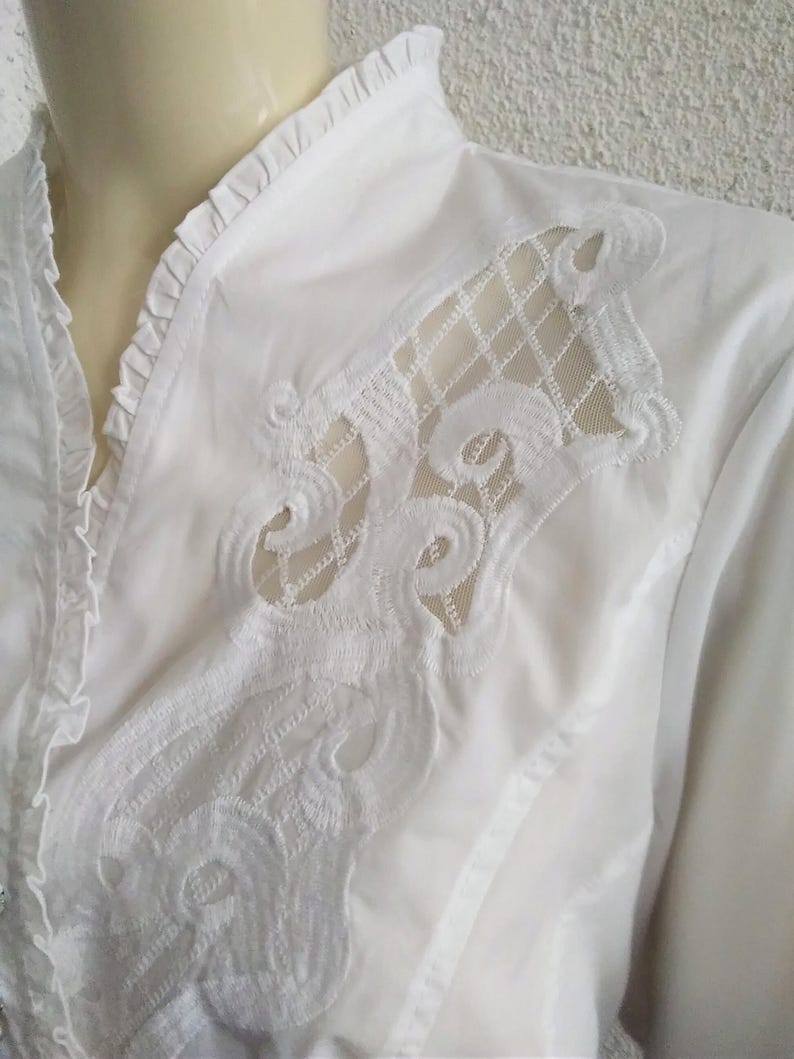 K&ouml;nnte beinhalten: Wei&szlig;e Bluse mit langen &Auml;rmeln, R&uuml;schenkragen und einer zarten Spitzenapplikation auf der Vorderseite.