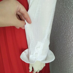 May include: White long-sleeved blouse with a textured fabric and a white button cuff. The blouse is tucked into a red pleated skirt.