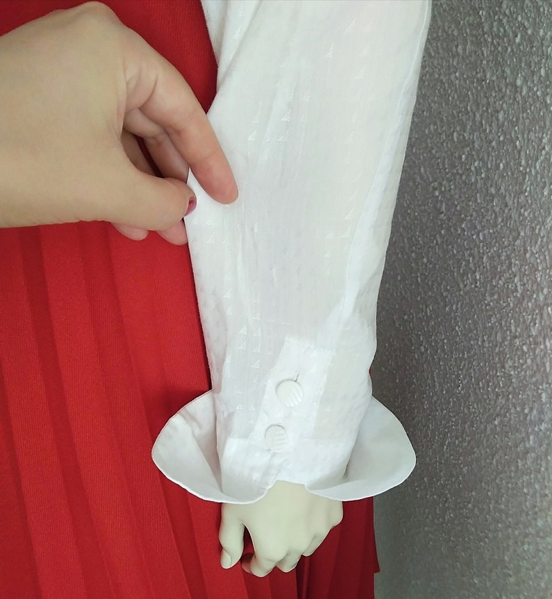 May include: White long-sleeved blouse with a textured fabric and a white button cuff. The blouse is tucked into a red pleated skirt.