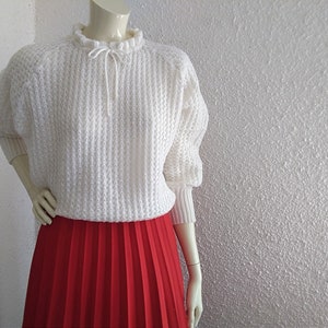 May include: White knitted sweater with a ruffled neckline and a tie at the neck. The sweater is worn over a red pleated skirt.