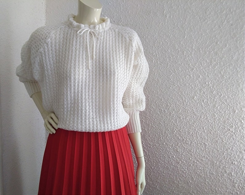 May include: White knitted sweater with a ruffled neckline and a tie at the neck. The sweater is worn over a red pleated skirt.