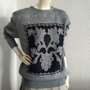 Op de afbeelding: Een grijze trui met een marineblauw en zilver bloemmotief. De trui heeft een ronde hals en lange mouwen. Zilveren versieringen zijn verspreid over de schouders en rond het bloemmotief.