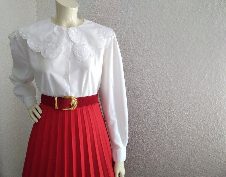 Puede incluir: Una blusa blanca con un cuello grande de encaje y mangas largas. La blusa se lleva con una falda plisada roja y un cintur&oacute;n dorado.