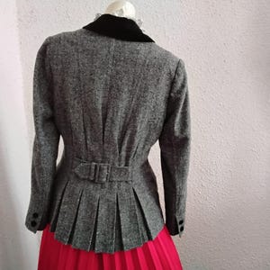 May include: A grey tweed jacket with a black velvet collar and a pleated peplum. The jacket has a belt at the waist and black buttons on the sleeves. The jacket is paired with a bright red skirt.